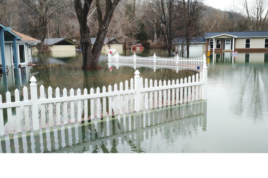 Flooded residential area with submerged yards, fences, and houses surrounded by water underscores the importance of flood preparedness for homeowners. Bare trees and an overcast sky loom in the background, painting a stark picture of nature’s power.
