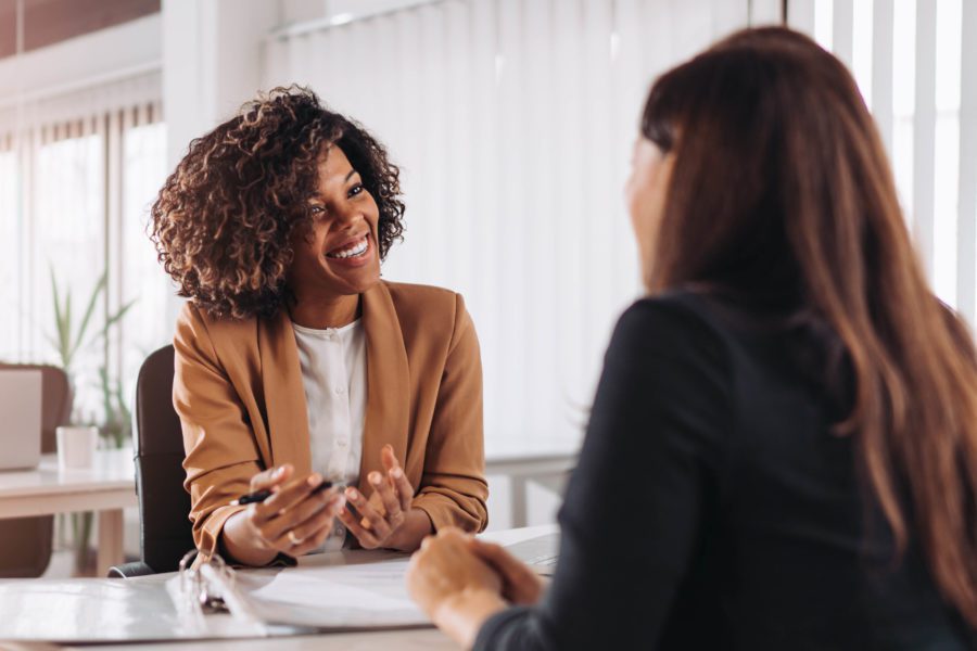 Two people are seated at a table in a bright office. One person in a brown blazer is smiling and gesturing, discussing HR outsourcing, while the other in a black jacket listens attentively.