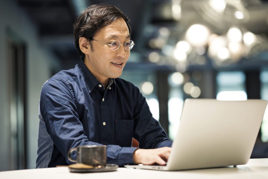 A man in glasses and a dark shirt is seated at a desk, focused on his laptop. A cup sits nearby. The blurred background with lights and office decor suggests he's deep into employee engagement surveys, measuring how connected colleagues feel.