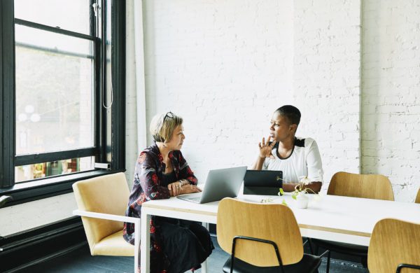 Two people sit at a white table in a modern office, each with a laptop. They're engaged in conversation, possibly discussing changing insurance brokers, surrounded by natural light from large windows.