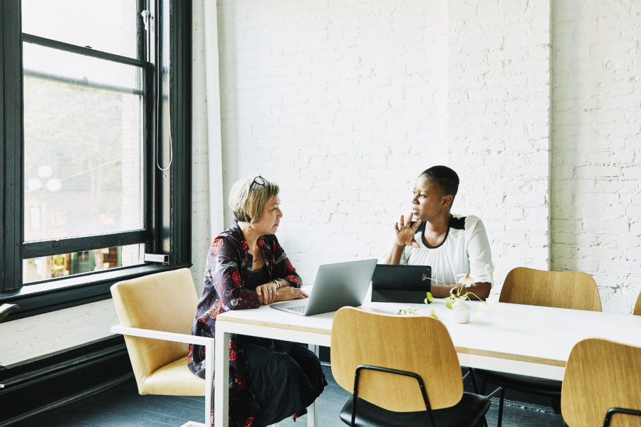 Two people sit at a white table in a modern office, each with a laptop. They're engaged in conversation, possibly discussing changing insurance brokers, surrounded by natural light from large windows.