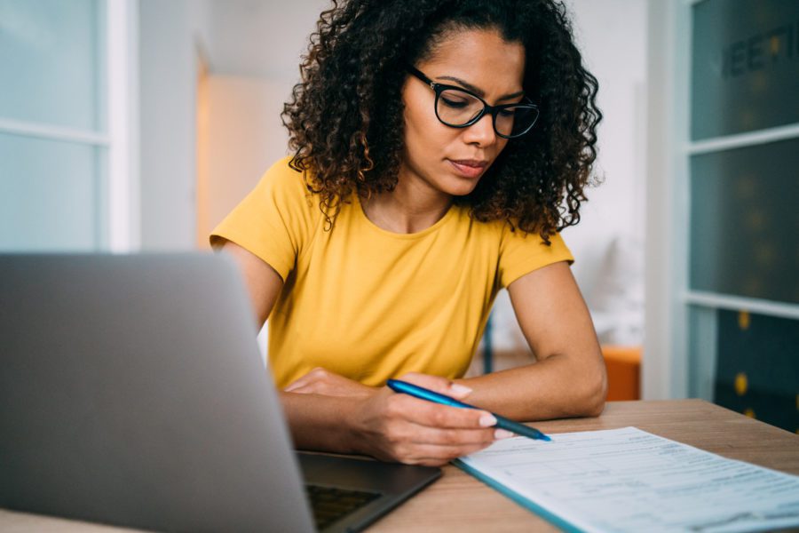 A person in a yellow shirt and glasses meticulously writes on a document at a desk with a laptop, likely reviewing details on excess and surplus insurance.