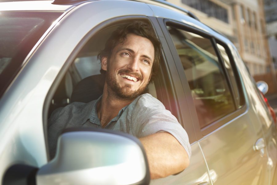 A man with a beard is smiling while sitting in the driver's seat of a silver car, one of the safest cars of 2022.