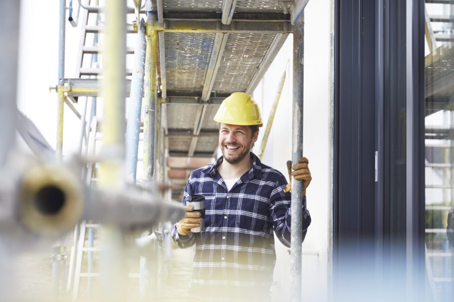 Man in a hard hat and plaid shirt smiles while holding a phone on scaffolding near building windows, confident in his team's bonding capacity to handle any project.