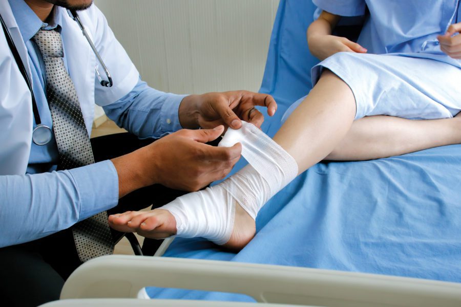 In a hospital room, a doctor bandages a patient's ankle as they sit on the bed in a blue gown, all while wondering if homeowners insurance might cover injury liability.