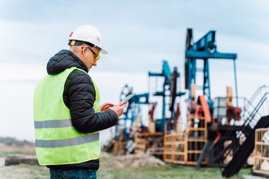 Professional worker man in helmet and protective glasses measuring and analyzing data on digital tablet on oil field near built construction