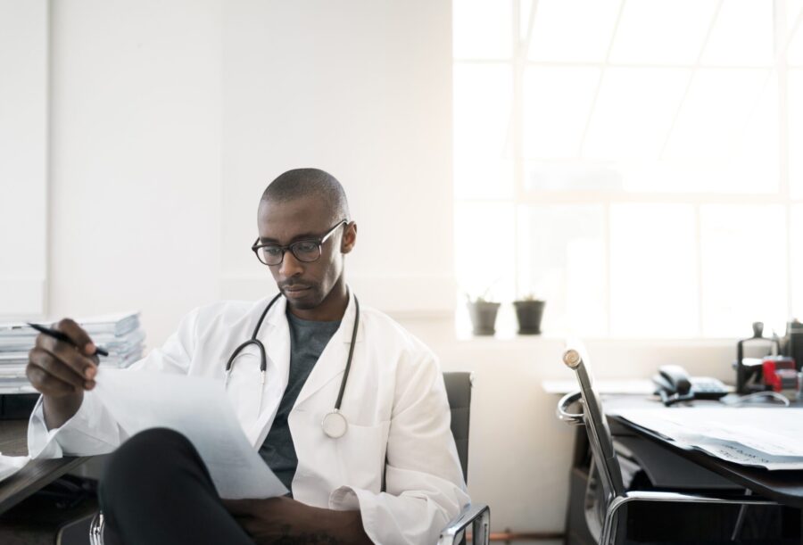 A photo of doctor reading document while sitting on chair. Male professional wearing lab coat and stethoscope. He is in office.