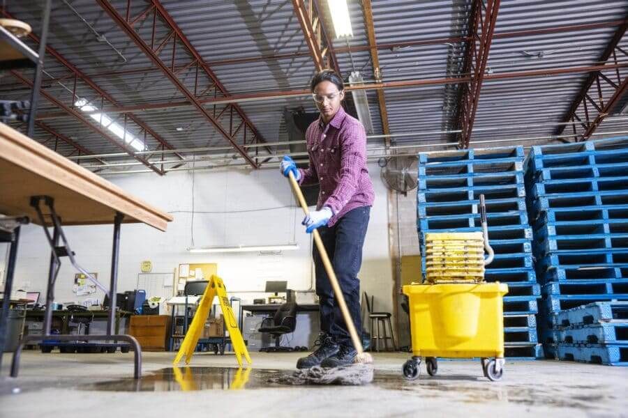 A male worker wearing work boots in a warehouse cleaning up a liquid spill on the floor.