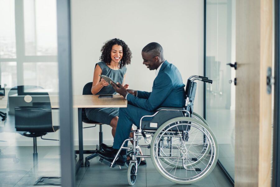 Shot of an african-american businessman in wheelchair having discussion with his female colleague in the board room. Photo of elegant businesswoman working with her disabled colleague in the office. Corporate business persons discussing new project and sharing ideas in the workplace.
