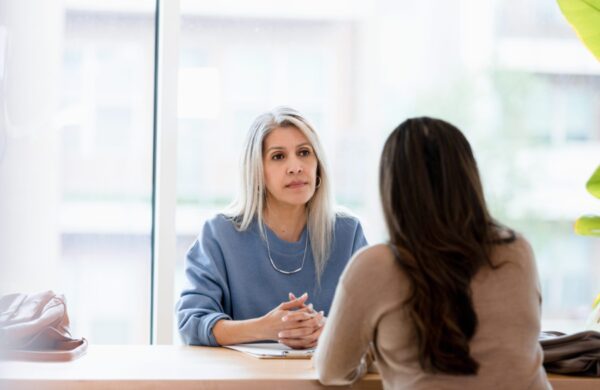 With her hands clasped in front of her, the mature adult businesswoman listens with a serious look on her face to the unrecognizable female client.