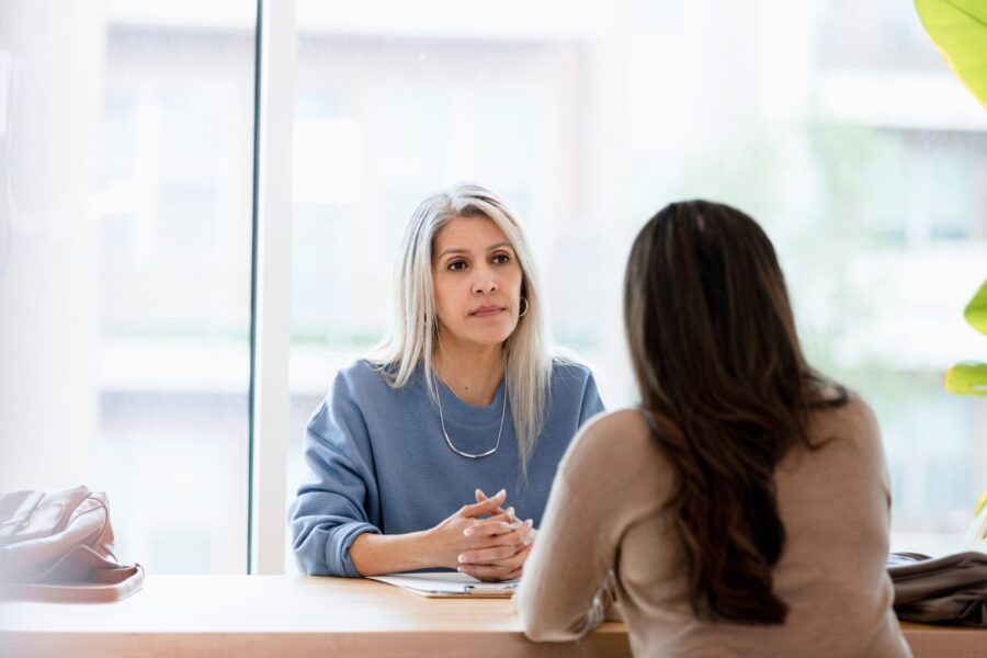 With her hands clasped in front of her, the mature adult businesswoman listens with a serious look on her face to the unrecognizable female client.