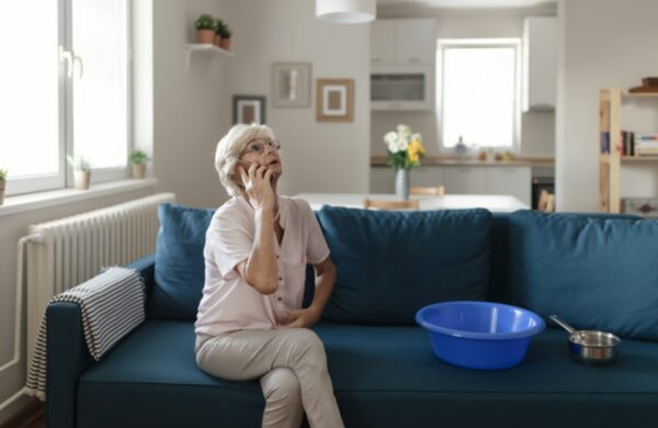 Shocked Senior Woman Calling Plumber While Collecting Water Leaking From Ceiling Using Utensil. Frustrated Elderly Woman Calling Insurance About Water Leaks in the Living Room at Home