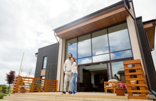 Happy young couple embracing standing outside big house on front porch, smiling owners enjoying luxury real estate wood and glass exterior design, buying home, mortgage loan, vacation rentals concept