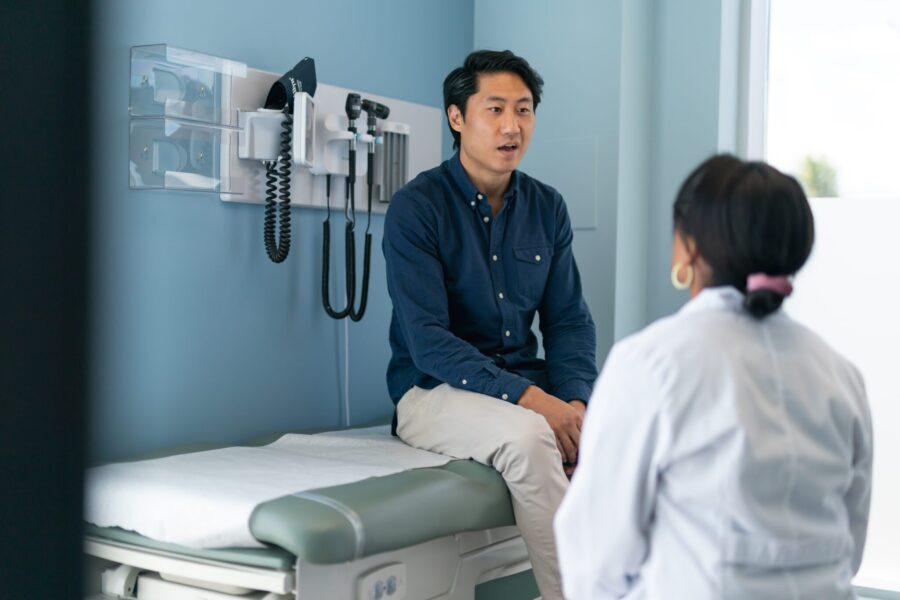 A Korean man is at a routine medical appointment. His doctor is a black woman. The patient is sitting on an examination table facing his doctor. The kind doctor is listening as he speaks.