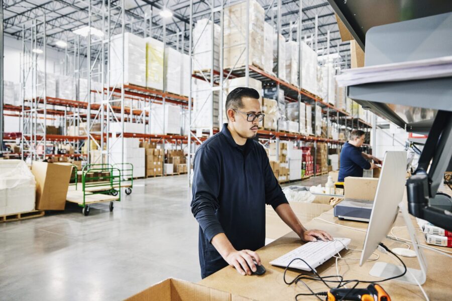 Medium wide shot of male warehouse worker checking orders at computer workstation in warehouse