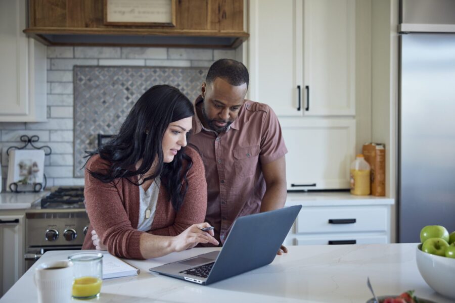 Caucasian woman and African American man sit at kitchen counter with breakfast working with pen, paper and laptop.