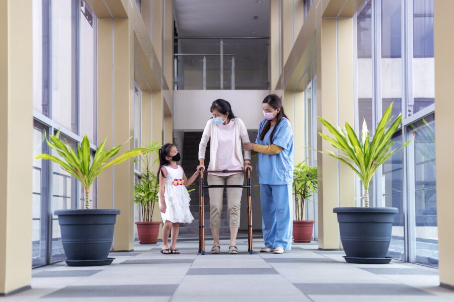 Senior Asian woman walking with mobility walker, assisted by physical therapist and grand daughter