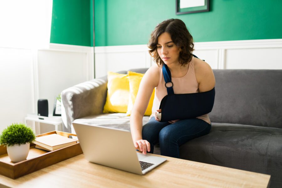 young woman with an arm sling typing on the laptop after a having an accident and a broken bone