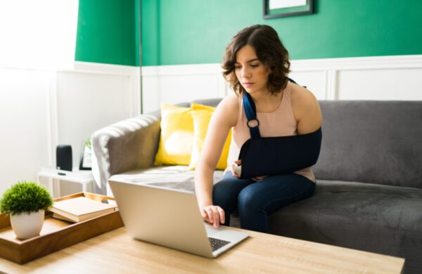 young woman with an arm sling typing on the laptop after a having an accident and a broken bone