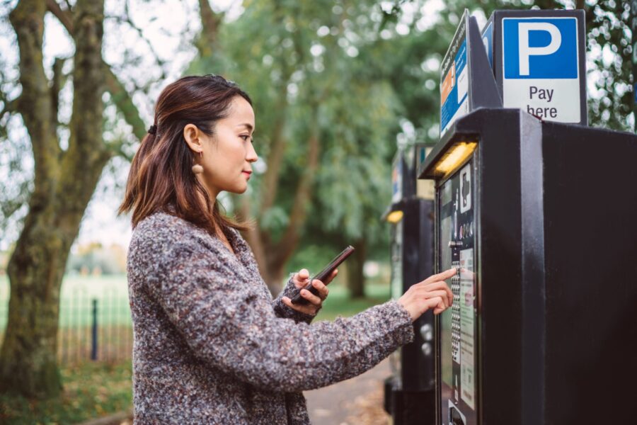 Young pretty Asian woman making contactless payment with smartphone at parking payment machine in city street.