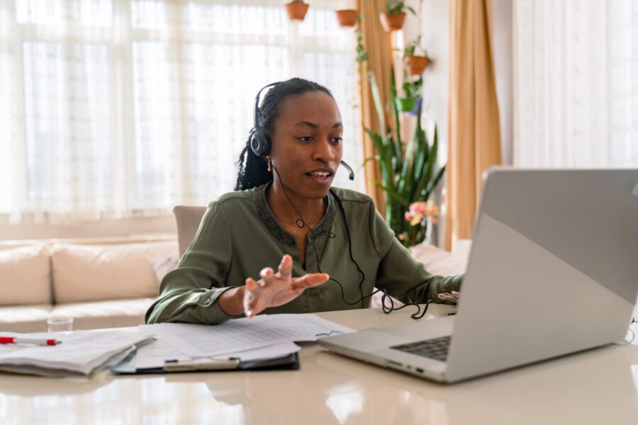 Confident black businesswoman wearing headphones using laptop, speaking, making video call, support service operator consulting client online, looking at computer screen, employee watching webinar