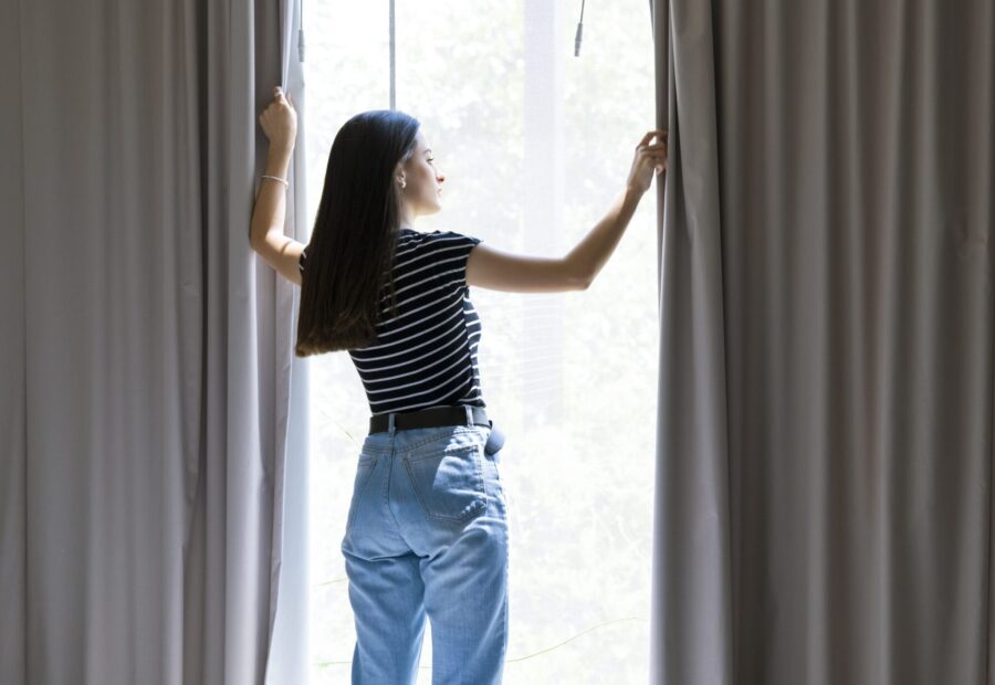 Rear view of young woman looking through window. Female is opening curtains at home. She is wearing casuals.