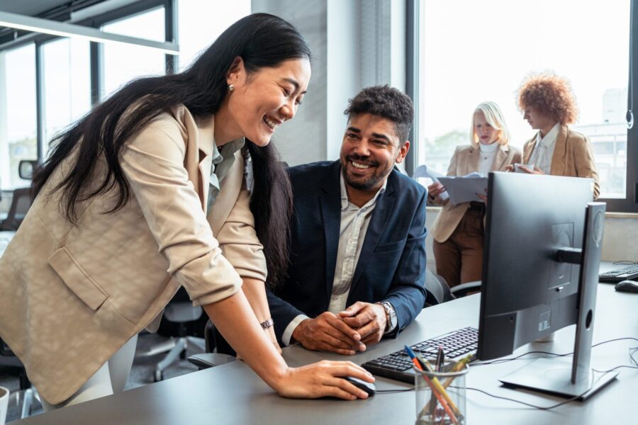 Confident female CEO of Asian ethnicity, assisting to her male manager of mixed-race ethnicity, about some work on the computer, while both talking and smiling