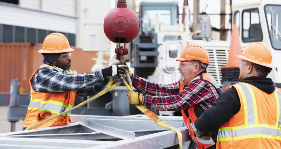 A multiracial group of three construction workers working together to prepare a large object to be hoisted with a mobile crane. They are attaching the lifting straps to a hook.