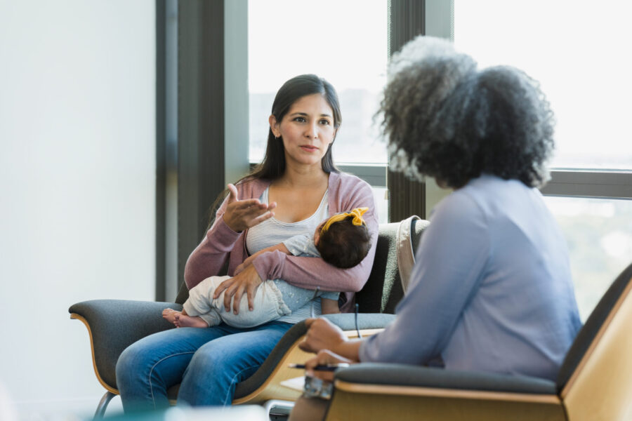 The mid adult woman with a baby in her arms gestures as she shares her struggles with her female therapist.