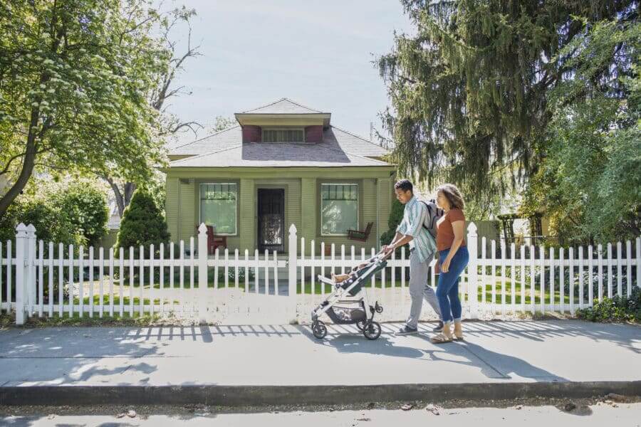 Young Mother And Father Walking With Baby Daughter In Stroller On Neighborhood Sidewalk