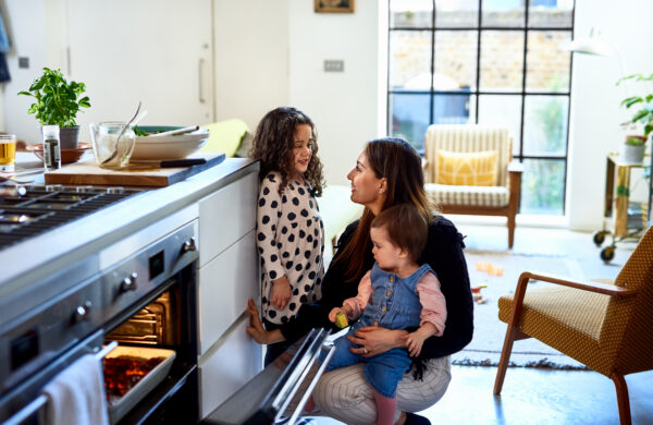 Mid adult woman opening hot oven and smiling at daughter, with toddler on knee