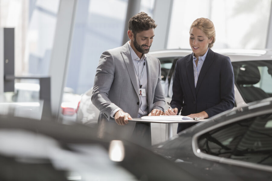 Car Salesman And Female Customer Reviewing Financial Contract Paperwork In Car Dealership Showroom