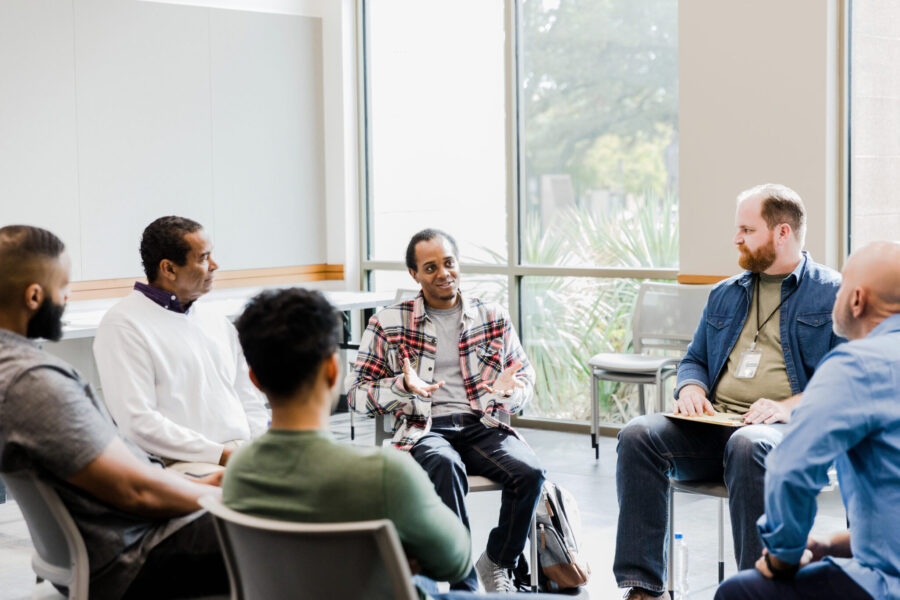 A multiracial group of men listen as the young adult man gestures and shares his struggles during a group therapy session.