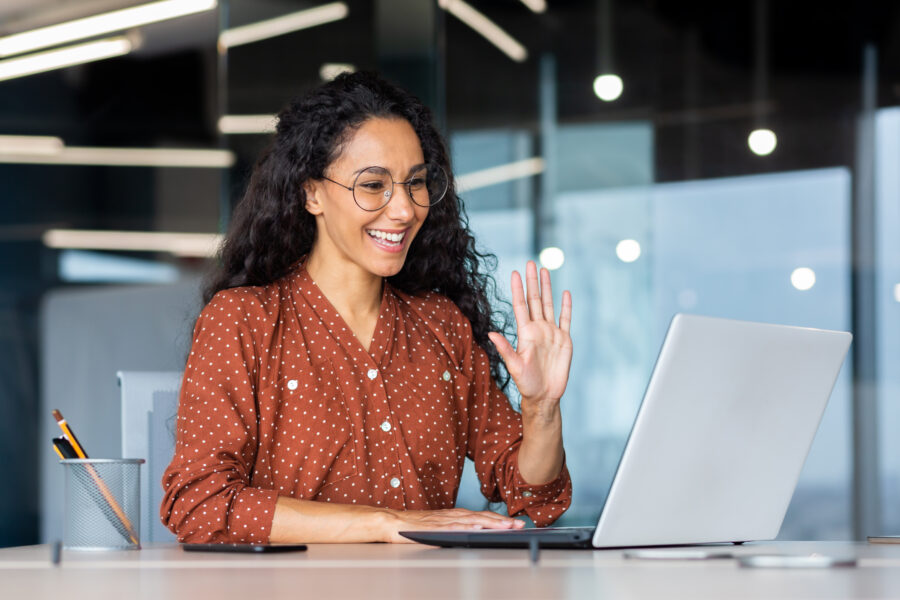 Successful and beautiful hispanic woman working inside modern office building, businesswoman using laptop for video call smiling and waving, greeting gesture, online conference with colleagues.