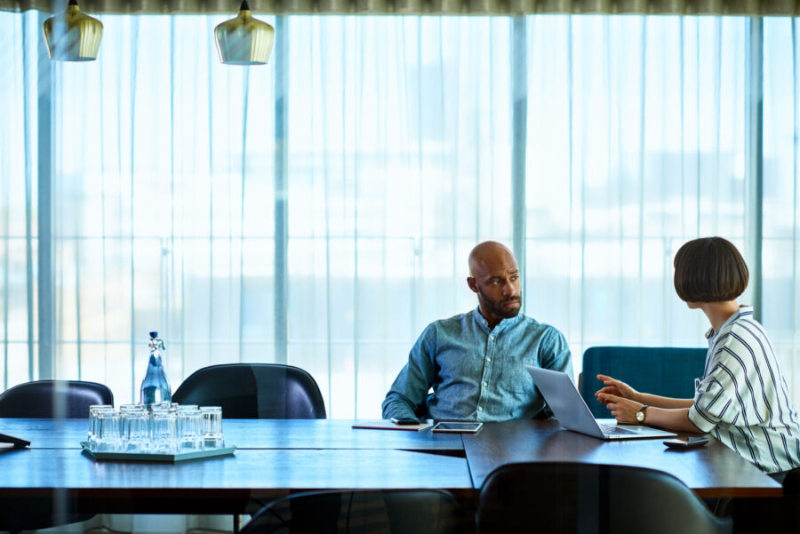 Latin American businesswoman gesturing towards businessman in modern office, using laptop and explaining, mid adult African man listening with serious expression