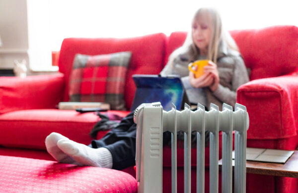 A retired senior woman in her 70s sits at home inside her cold house in winter. She is warming herself next to an electric heater while sitting on the sofa while she uses a digital tablet.