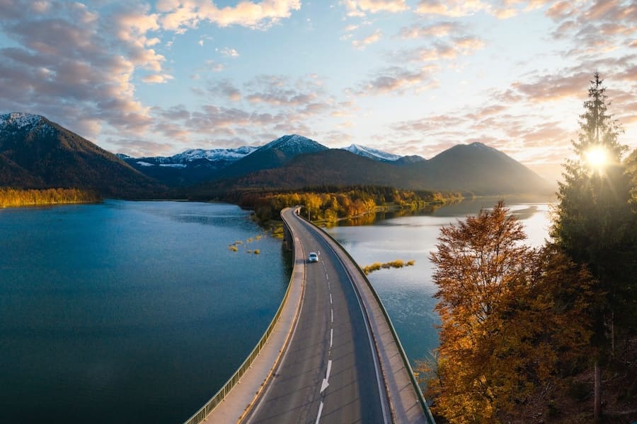Car driving over a bridge with a lake and mountain range backdrop.
