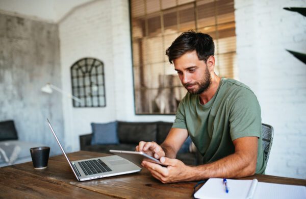 Young handsome man working from home, focused on apps and software on laptop and digital tablet