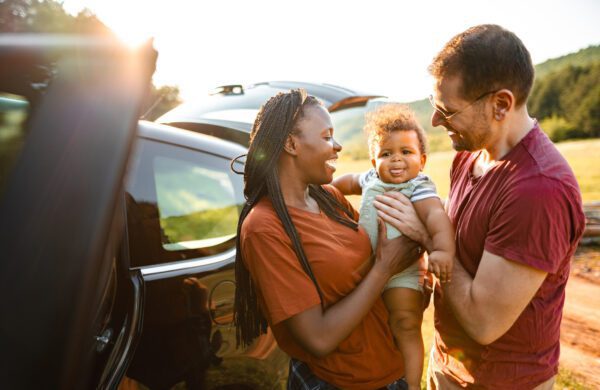 Smiling parents holding their baby near a car in a sunny outdoor setting