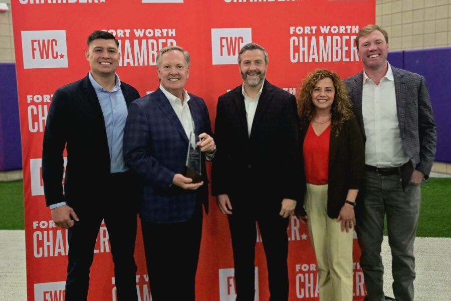 Five people stand together, one proudly holding the Spirit of Enterprise Award, against a red backdrop adorned with "Fort Worth Chamber" logos at the Annual Meeting.