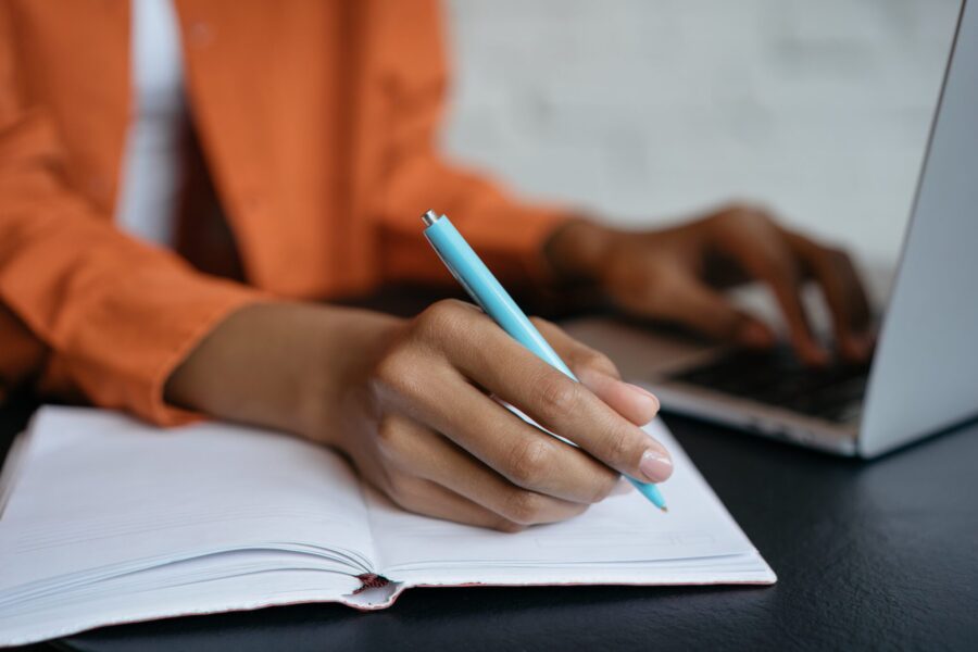 A HR professional writes in a notebook with a pen while typing on a laptop, writing a description for a job posting