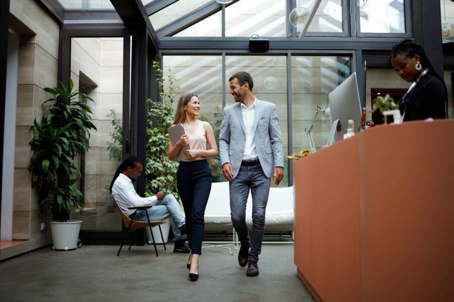 Two people walk together in a modern office space with large windows and plants, embracing sustainability in the workplace. Another person sits in the background, while a woman stands at a reception desk.