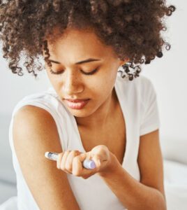 Person with curly hair in a white shirt carefully administers an injection into their arm using a syringe pen.