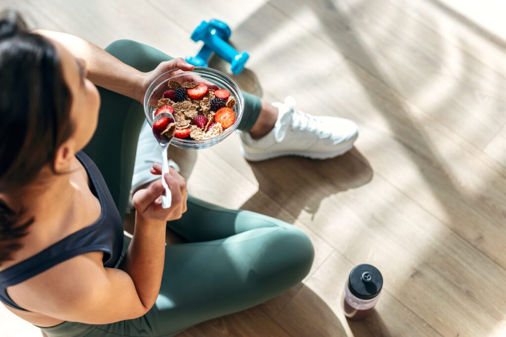 Athletic woman eating a bowl of healthy cereal and nutritional oats with fruit while sitting on floor after exercising