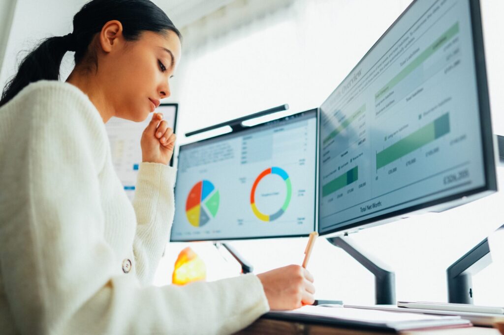 A woman sits at a desk analyzing data on dual monitors displaying charts and graphs while taking notes on a notepad
