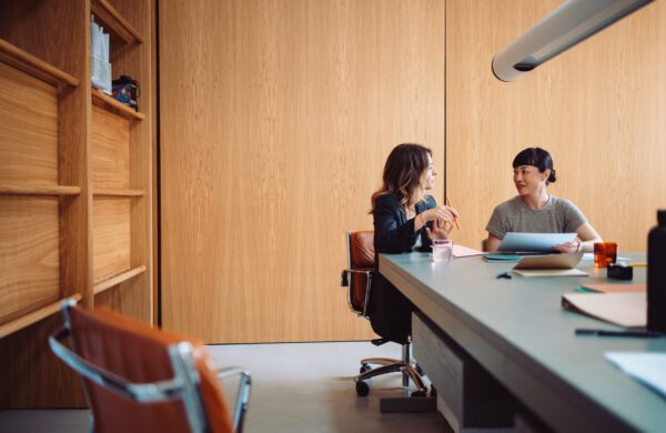 Two businesswomen engaged in a discussion at an office table, collaborating on a project with documents and laptops