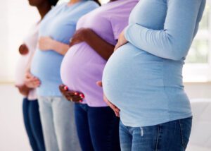 Four pregnant women standing in a row, each holding their baby bump, wear colorful long-sleeve tops. Their faces are not visible. Perfect for illustrating maternity policy changes in the April 2025 HR Update.