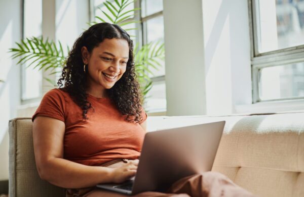 Young woman sitting on a sofa in the living room with her laptop computer scrolling on the internet, smiling