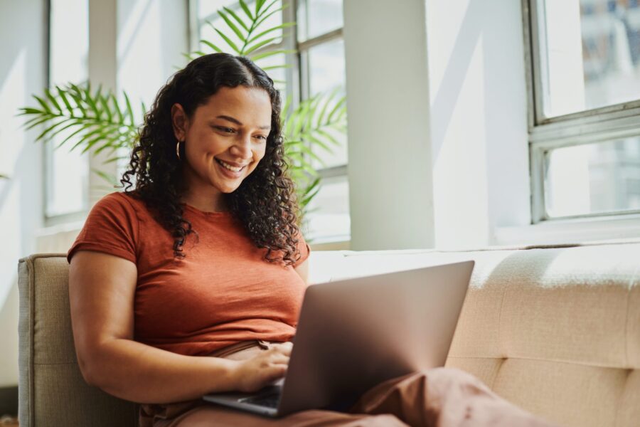 Young woman sitting on a sofa in the living room with her laptop computer scrolling on the internet, smiling